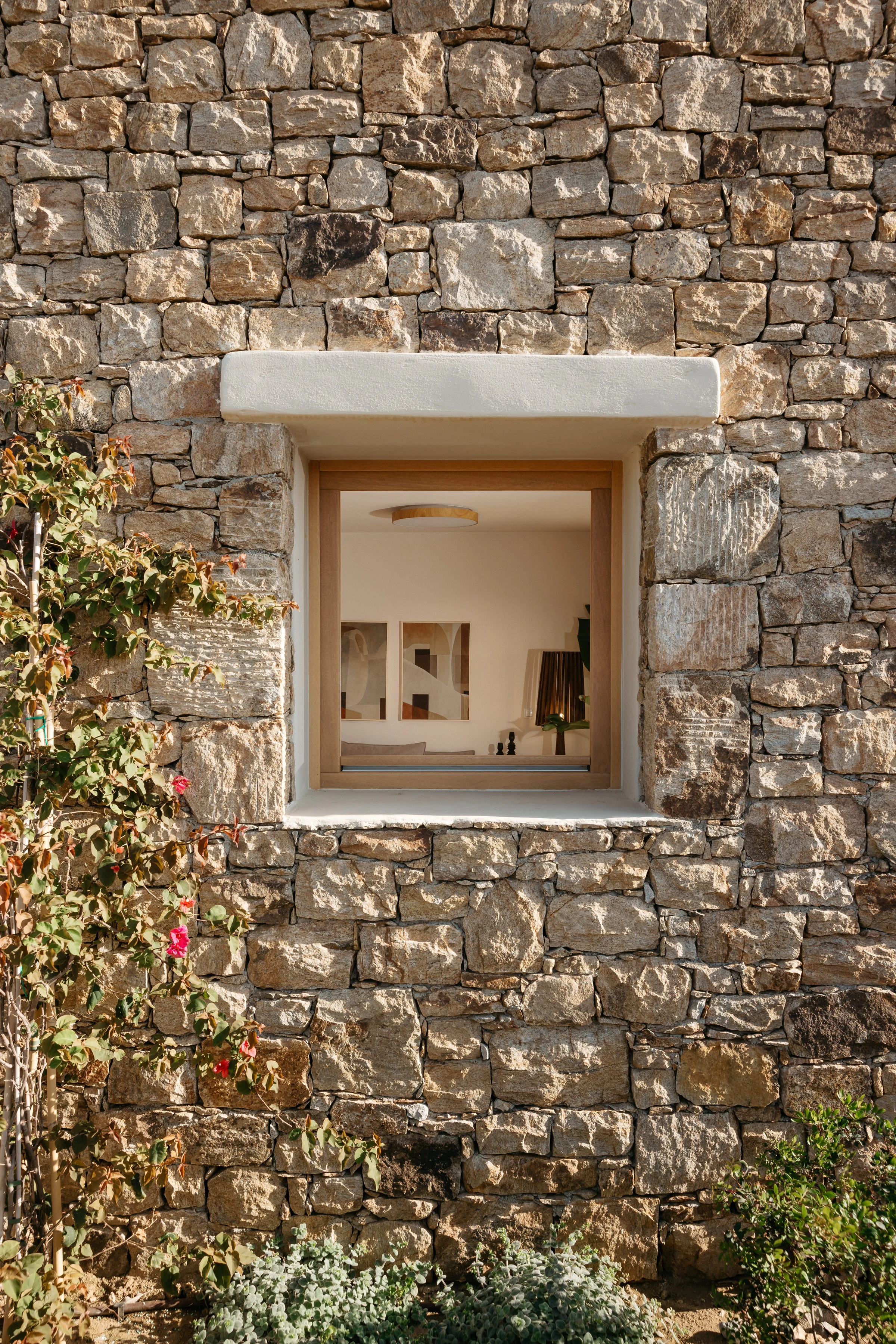 Dry-stone facade with a timber-framed window into the living room, bougainvillea climbing the wall