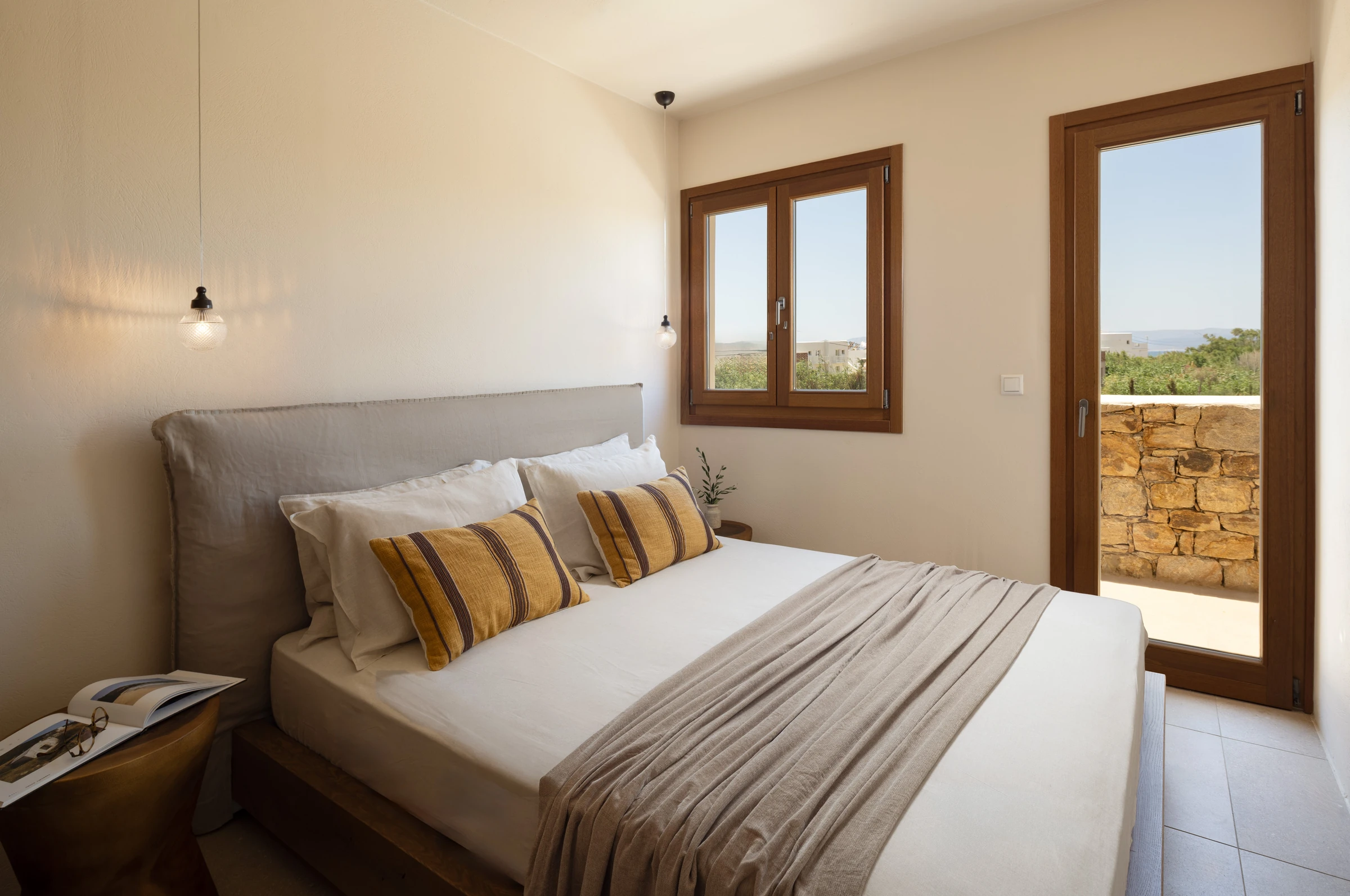Double bed with grey linen headboard, ochre striped cushions, corner window and terrace door