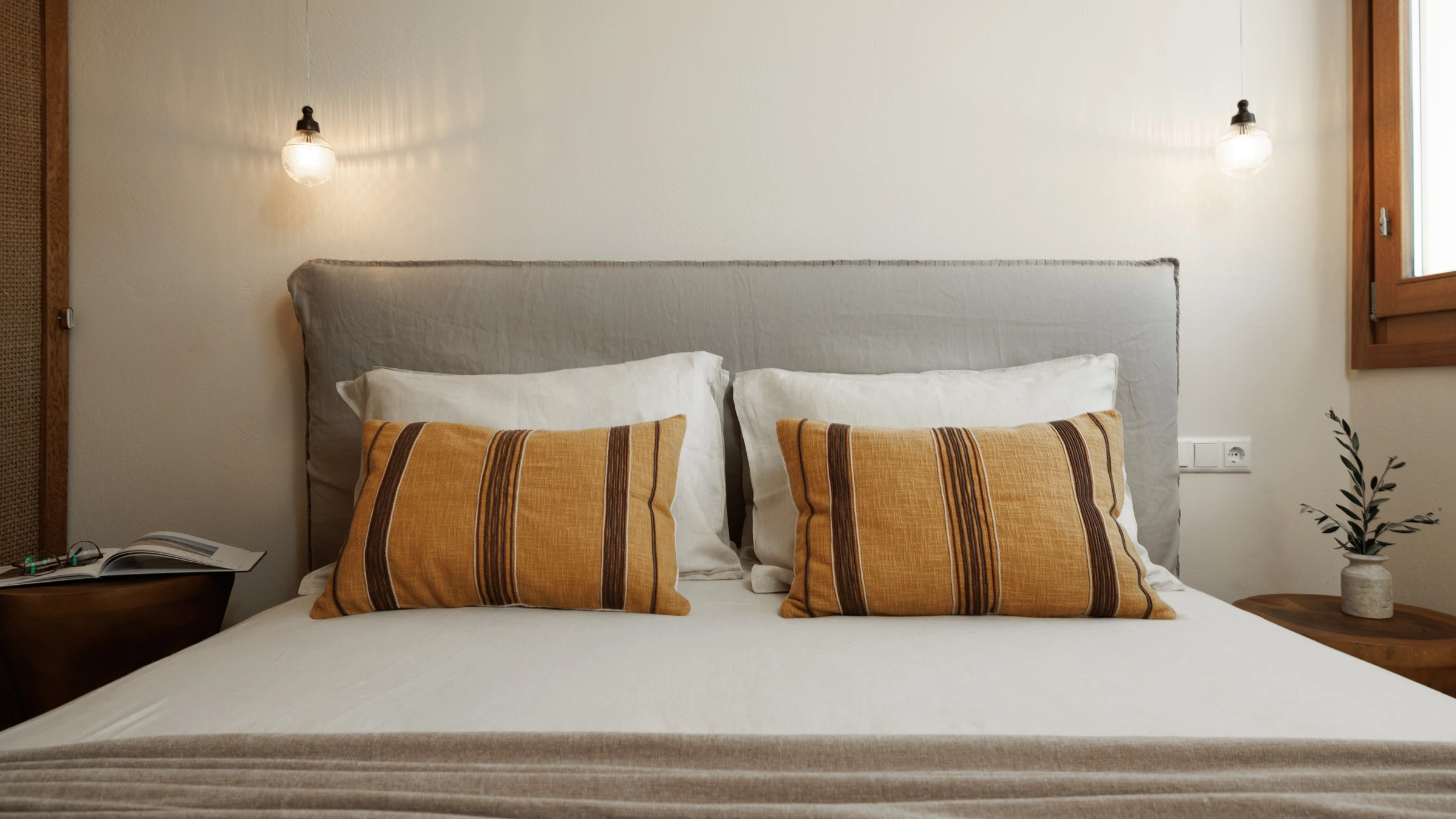 The bed head-on, ochre striped cushions and twin glass pendants against a grey linen headboard