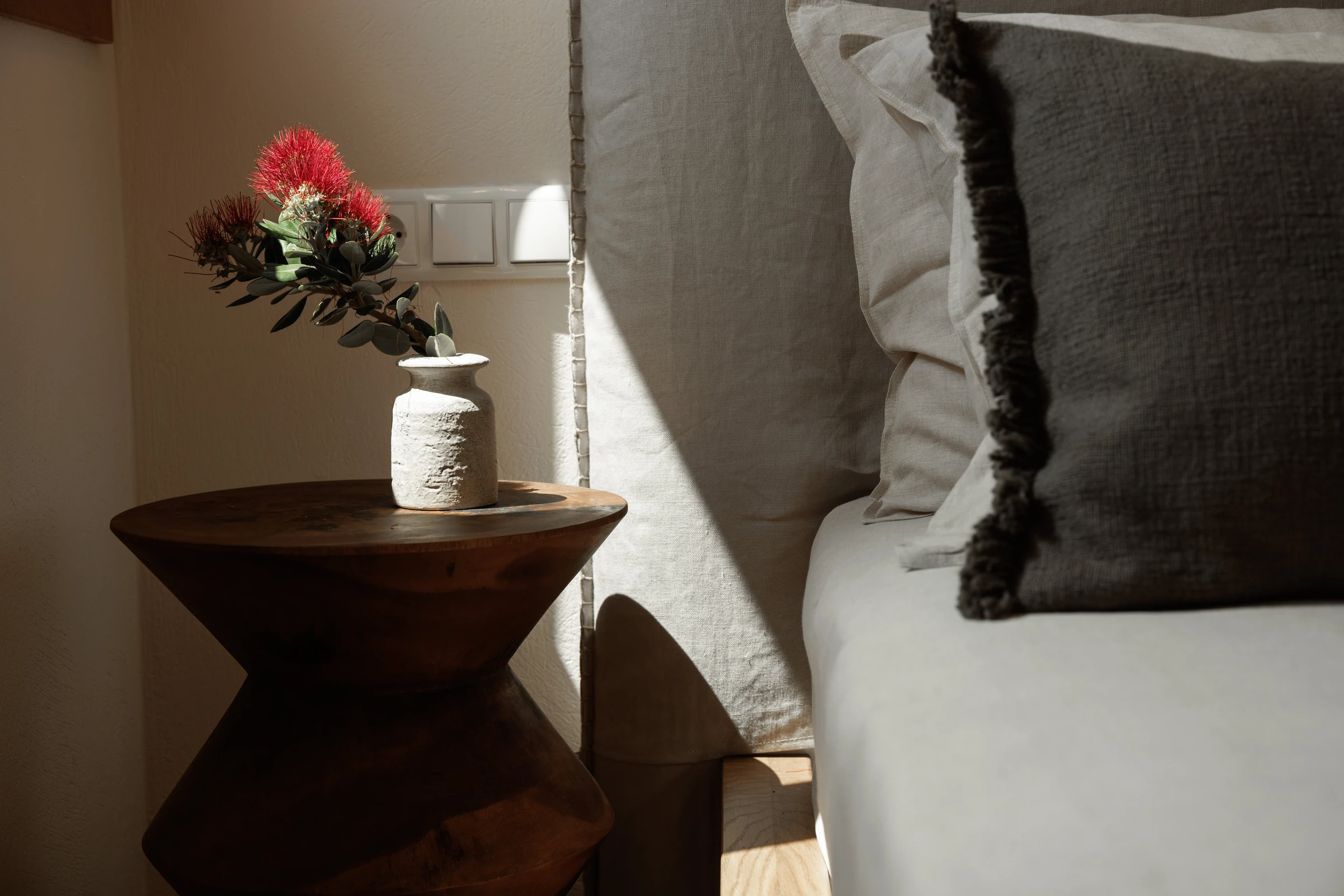 A crimson bottlebrush sprig in a white ceramic vase on the hourglass side table, grey fringed cushion beyond