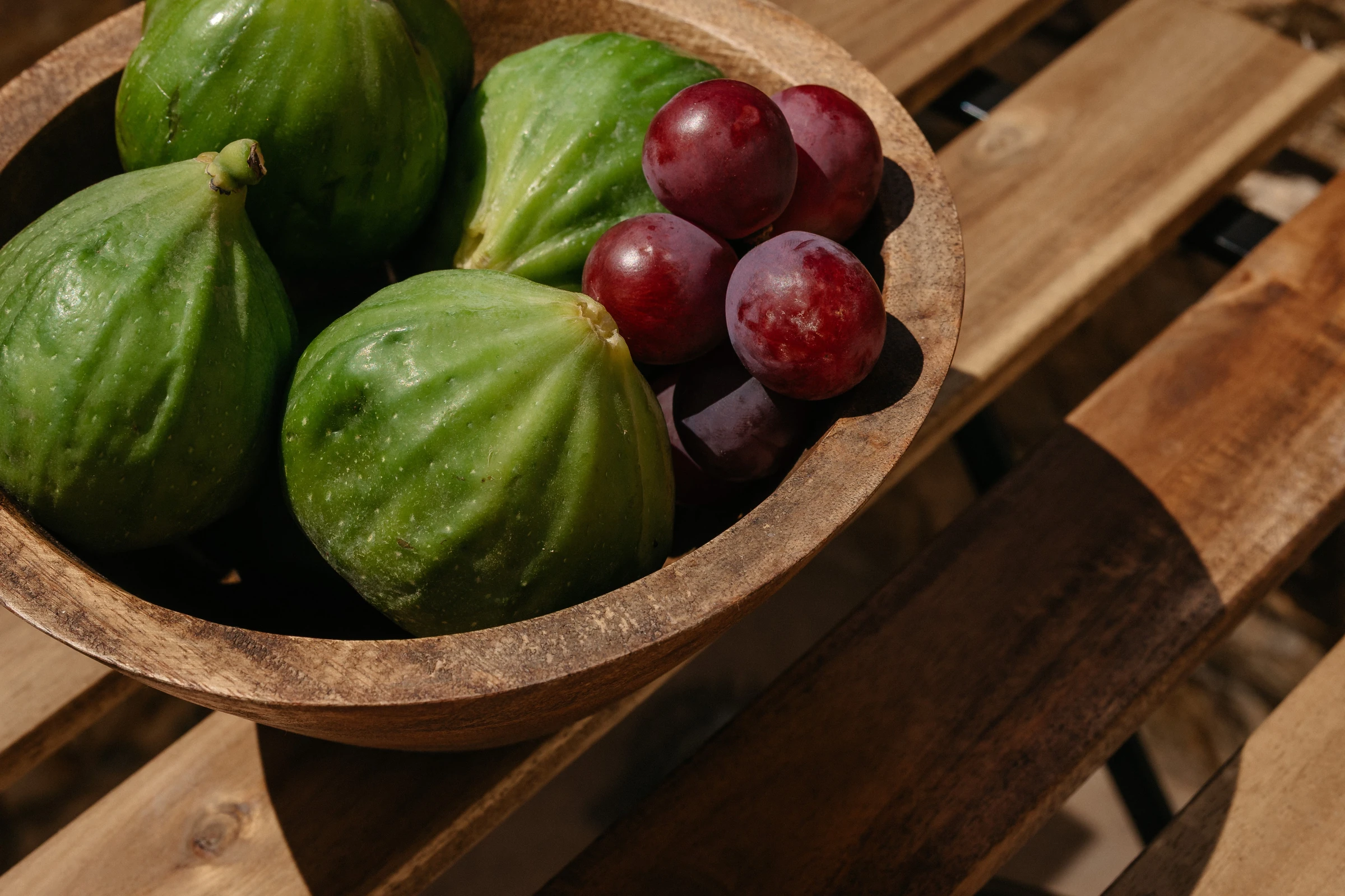 A carved wooden bowl of green figs and red grapes in the sun